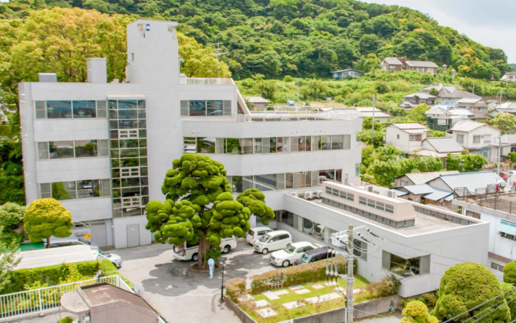 緑に囲まれた丘陵地に立つ白い現代建築の介護施設外観。駐車場と庭園が整備されている。